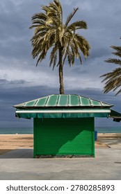 Empty sandy beach scene featuring a closed green metal kiosk and palm trees in Badalona, ideal for winter tourism backgrounds.
