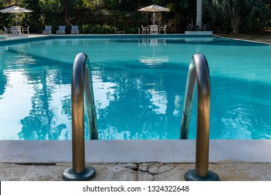 Empty Scene Clean Outdoor Swimming Pool With Chairs, Tables, Large Patio Umbrellas. Cracks On Pool Deck Surface Near Mounted Metal Handrails/ Hand Rails. No People Taking A Fun Swim. 