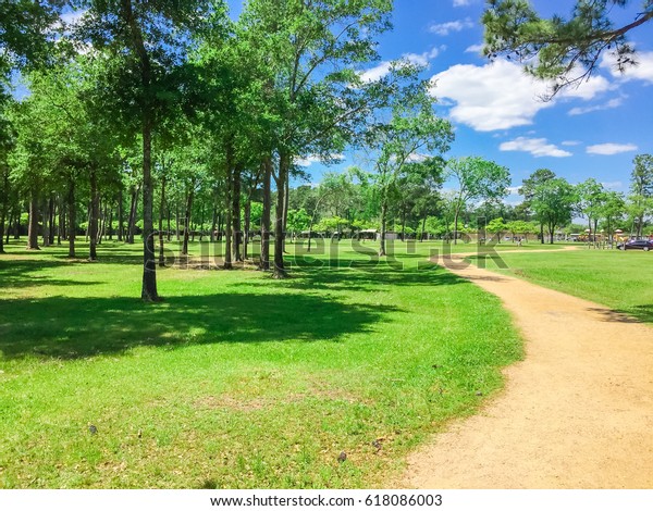 Empty s-curved trail through grassy urban park
with row of pine, oak trees  during daytime, blue cloud sky in
Humble, Texas, US. Unidentified cars are visible at the parking lot
in the far left corner