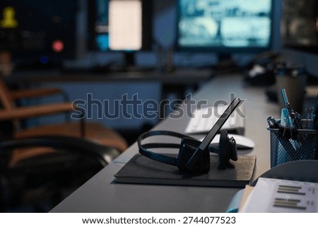 Empty security office desk featuring wireless headset resting on notebook, computer keyboard and monitor in background, pen holder with pens and documents visible, modern workspace setting