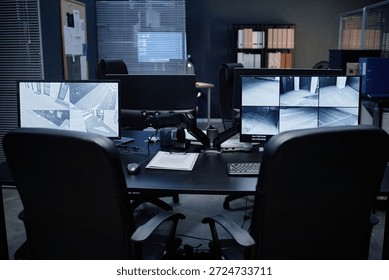 Empty security office showing two computer monitors displaying multiple surveillance camera feeds, office chairs positioned in front of desk, various documents and equipment arranged on tabletop
