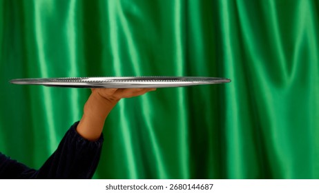 Empty silver serving tray held by bare hand on green curtain background. Concept of service, catering visuals, branding presentation, product placement, and promotional design. Negative space for ads