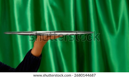 Empty silver serving tray held by bare hand on green curtain background. Concept of service, catering visuals, branding presentation, product placement, and promotional design. Negative space for ads