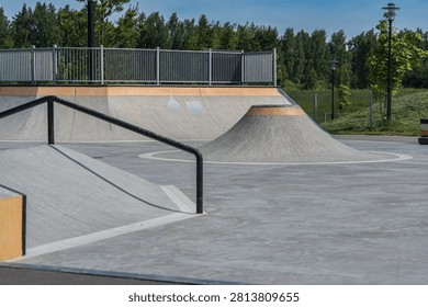  Empty skate park showcasing smooth concrete ramps, a metal rail, and a curved mound. Ideal for skateboarders and BMX riders. Surrounded by greenery, perfect for leisure sports. Empty Skate Park with Concrete Ramps and Rails
