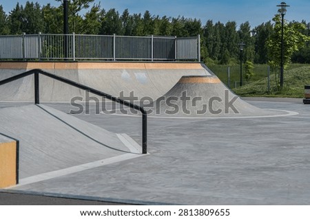  Empty skate park showcasing smooth concrete ramps, a metal rail, and a curved mound. Ideal for skateboarders and BMX riders. Surrounded by greenery, perfect for leisure sports. Empty Skate Park with Concrete Ramps and Rails
