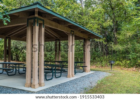 Empty small picnic pavilion with several tables on a cement pad with a grill alongside in the woodlands at the park