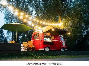 Empty Small Street Food Track With An Illuminations Standing In The Deserted Night Park. Near Food Trailer Are Folding Tables, Chairs And Umbrella
