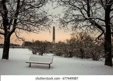 An empty snow covered bench sits in the middle of Vigeland Park on a cold afternoon in Oslo Norway.  The sun sets as two trees frame a sculpture in the center of the park.