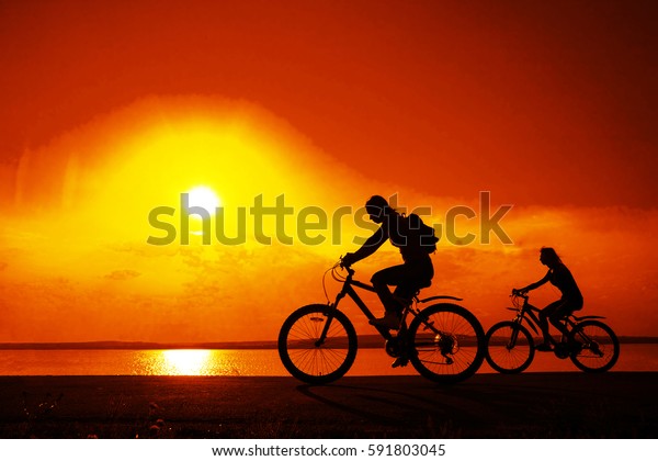 Empty Space. Sporty friends on bicycle on sunset.\
Silhouette couple cyclist go along coast. Sport in Nature\
background. Group of people two mountain biker in sunrise with\
reflection sun in water
