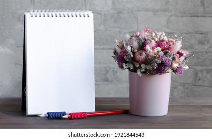 An empty spiral notebook, two pencils, and a delicate bouquet of flowers in a pink vase on a wooden surface. Close-up, brick wall in the background. Ready-made layout for advertising, ads.