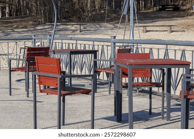 An empty square with several red tables and chairs placed on a concrete base, trees visible in the background.