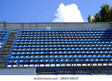 Empty stadium stands featuring rows of blue and white plastic seats under a clear blue sky. The image captures the stillness and symmetry of the seating arrangement. 