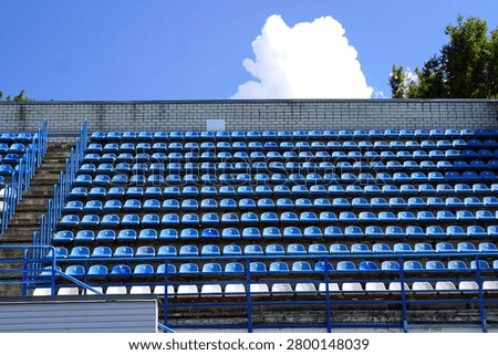 Empty stadium stands featuring rows of blue and white plastic seats under a clear blue sky. The image captures the stillness and symmetry of the seating arrangement. 