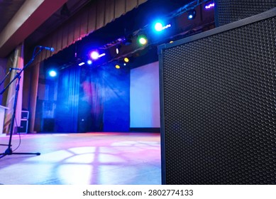 Empty stage is set with bright colored lights and a large black speaker on the foreground. Purple and blue lighting fills the curtains and projection screen. Microphone stand waits near the speaker.