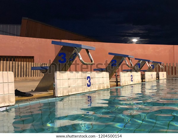 Empty starting block of\
outdoor swimming pool at night, dark night sky with the moon, low\
angle view.