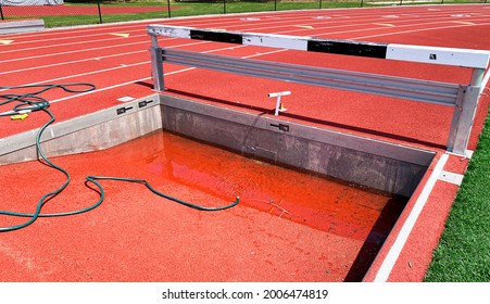 Empty Steeplechase Pit Being Filled Up With Water On A Track From A Green Hose For For Runners To Compete.