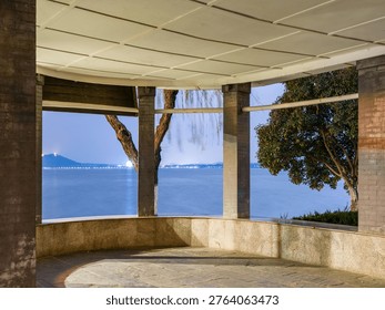 Empty stone observation deck and pavilion framing a beautiful view of a calm blue lake and distant glowing city skyline at twilight. The scene represents peaceful viewpoints, structural framing, quiet contemplation, and scenic waterfront landscapes.