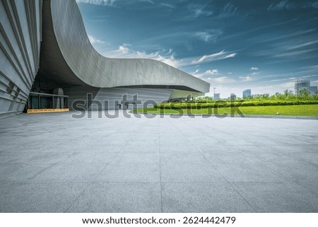 Empty stone square floor in front of a modern building with green lawn and distant cityscape under a blue sky.