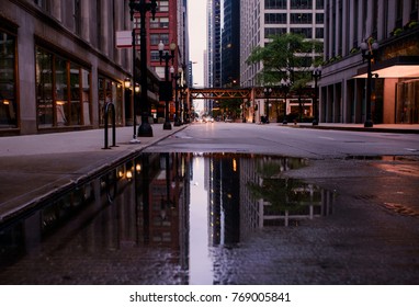 An empty street in downtown Chicago  is filled with a  large puddle. Inside the puddle, the buildings and lights reflect off the water. In the distance is the city traintracks. 