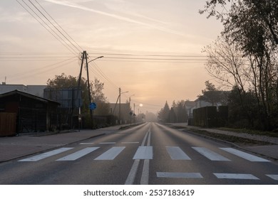 Empty suburban street at dawn with pedestrian crosswalk under soft, misty sunrise, surrounded by greenery and quiet residential homes. Concept of tranquil city living and early morning solitude.  - Powered by Shutterstock