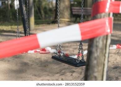 An empty swing dangles behind red and white cordon tape, its chains still against a closed sandy playground.