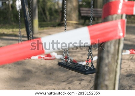 An empty swing dangles behind red and white cordon tape, its chains still against a closed sandy playground.