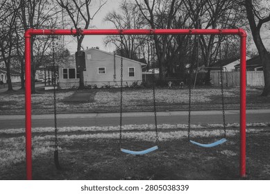 An empty swing set with a vibrant red frame and blue seats stands still in a quiet playground, set against the backdrop of a muted, grayscale residential neighborhood, creating a melancholic sense of solitude and the passage of time.