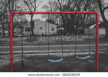 An empty swing set with a vibrant red frame and blue seats stands still in a quiet playground, set against the backdrop of a muted, grayscale residential neighborhood, creating a melancholic sense of solitude and the passage of time.