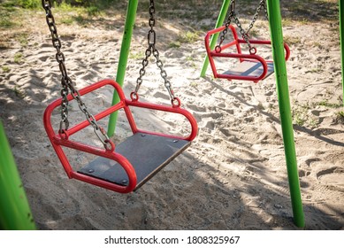 Empty Swing-set On Playground Because Of COVID Lockdown. Sand Underneath The Swings And A Grassy Area, Then Many Trees Behind. Good Summer Camp Image.