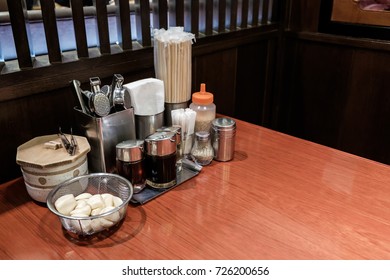 Empty Table In Japanese Ramen Shop With Seasoning And Condiment On Table. Chopsticks, Pepper Bottle, Sesame, Garlic, Soy Sauce Bottle On Wooden Table.