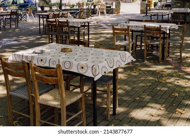 Empty Tavern Outdoors Seating Area With Wooden Chairs And Tables On A Village Square In Greece.