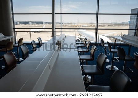 Empty terminal lounge with tables and chairs near large windows facing aircraft parking area. Convenient workstations with power outlets, hotspots and free WiFi access for travelers passengers