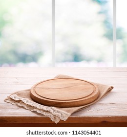 Empty textured wooden pizza board with napkin on the table and kitchen window blurred background. Top view mock up. Selective focus.