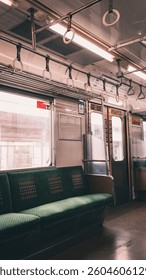 An empty train car interior is depicted with green cushioned seating, overhead fluorescent lighting, and numerous hand straps, suggesting a moment between passenger occupancy.