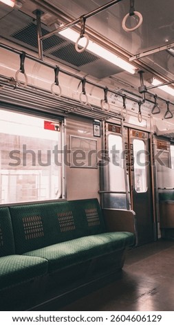 An empty train car interior is depicted with green cushioned seating, overhead fluorescent lighting, and numerous hand straps, suggesting a moment between passenger occupancy.