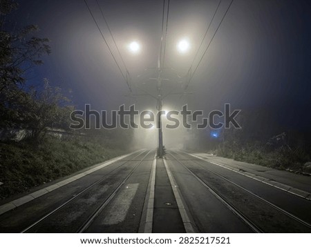 Empty tram tracks stretch into dense fog at night under bright streetlights, creating a quiet urban transport scene with rails disappearing into the distance.