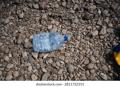 Empty translucent plastic water jug with blue cap lying on rounded river pebbles and stones, close-up view showing mixed sizes and colors of beach cobbles, natural daylight highlighting textures