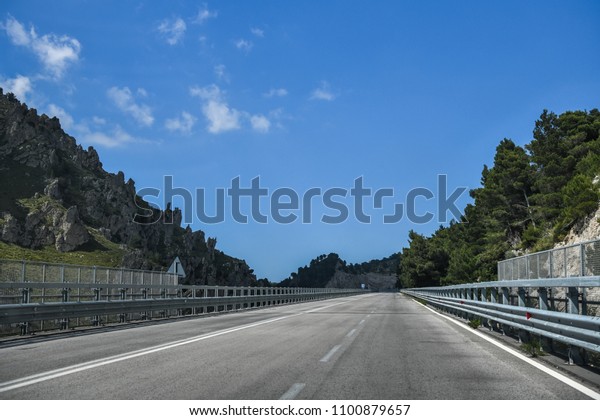 Empty two way tarmac\
road with guard rails surrounded by beautiful green trees and\
vegetation. Blue sky. 
