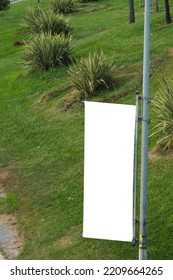 Empty Vertical Cloth Banner On A Street Lamp And Blurred Garden Background. You Can Change Easily Design Of The Billboard.