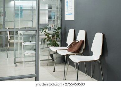 Empty waiting area featuring row of chairs against dark wall, brown leather handbag resting on one chair, glass partition revealing office space with potted plant and documents