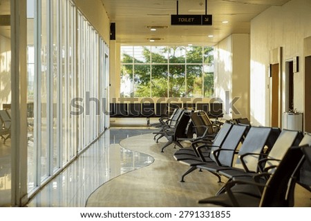 Empty waiting area in a terminal with rows of seats next to large glass windows filled with bright natural sunlight.