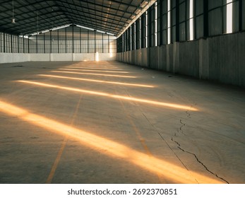 Empty warehouse interior with warm sunlight streaming through side windows, creating golden diagonal light streaks across the concrete floor, highlighting lines, symmetry, and industrial calm.