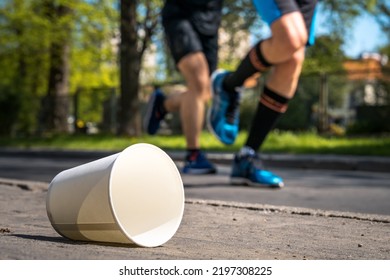 Empty Water Cup Thrown On The Road. Littered Marathon Run With Runners In The Background