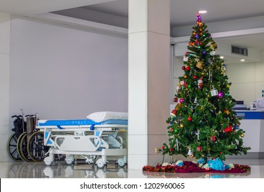 A Empty Wheelchair And Wheel Bed For Patient With Christmas Tree At Hallway Of Hospital. Christmas Time At The Clinic.