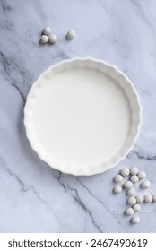 empty white ceramic pie plate with reusable ceramic pie weights on white marble background.
