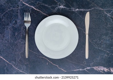 An Empty White Ceramic Plate With A Fork And A Table Knife On A Dark Marble Countertop, Top View