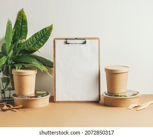 Empty White Note Board, Delivery Food In Sustainable Food Containers And Leaves On Brown Table At White Wall Background. Modern Office Still Life With Lunch. Front View With Copy Space.