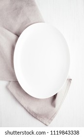 Empty White Oval Plate On Wooden Table With Linen Napkin. Overhead View.