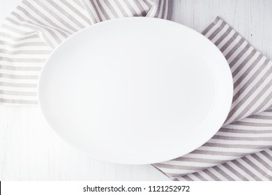 Empty White Oval Plate On Wooden Table With Striped Linen Napkin. Overhead View.