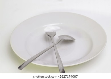 Empty white plate with crossed spoon and fork on a clean surface. Minimalist dining concept, symbolizing hunger, mealtime, or food preparation. Clean eating tableware setup.